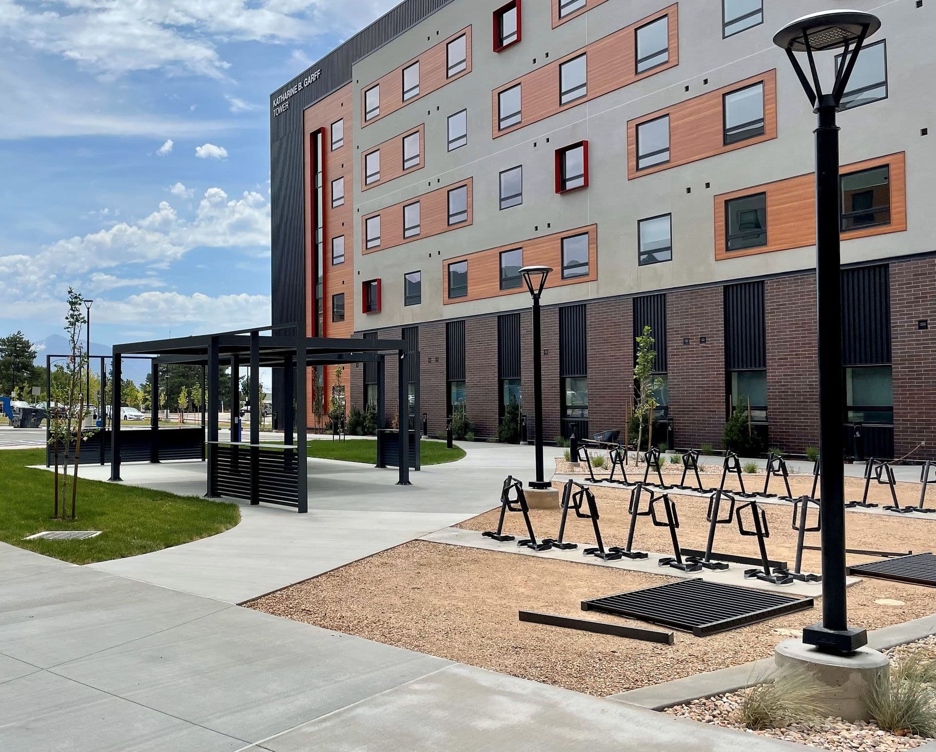 Modern urban setting with a multi-story building featuring orange accents, bike racks, a small pavilion, and street lamps under a blue sky.