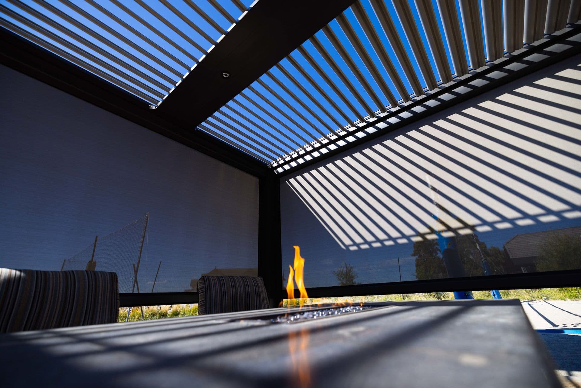Modern patio with a slatted pergola casting striped shadows. A small flame flickers on a table, creating a relaxed, contemporary atmosphere.