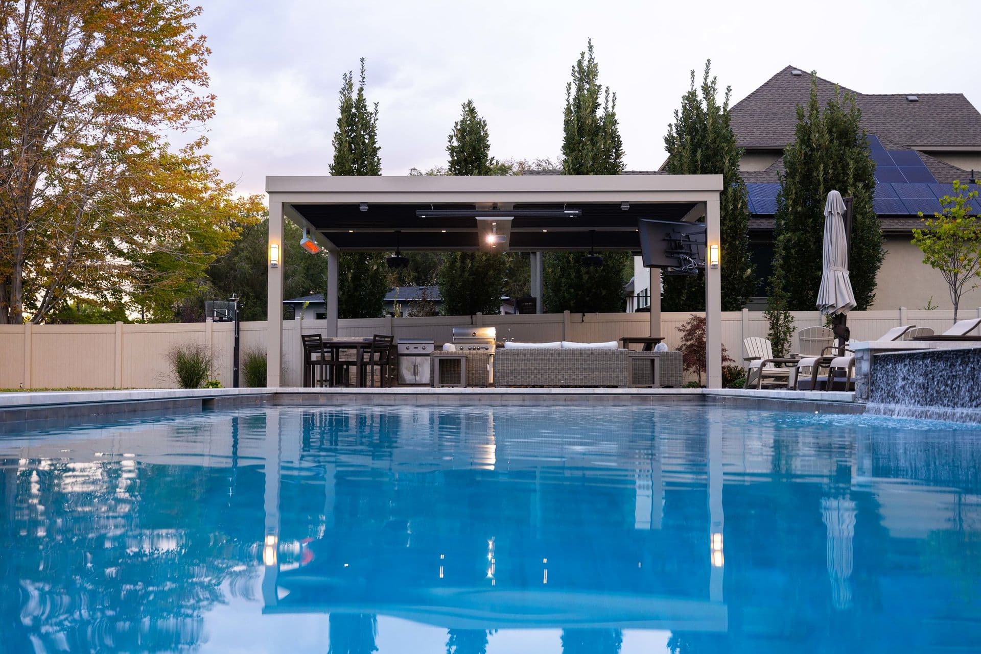 Modern backyard with a clear blue pool, a covered patio featuring seating, a grill, and trees in the background under a cloudy sky at dusk.