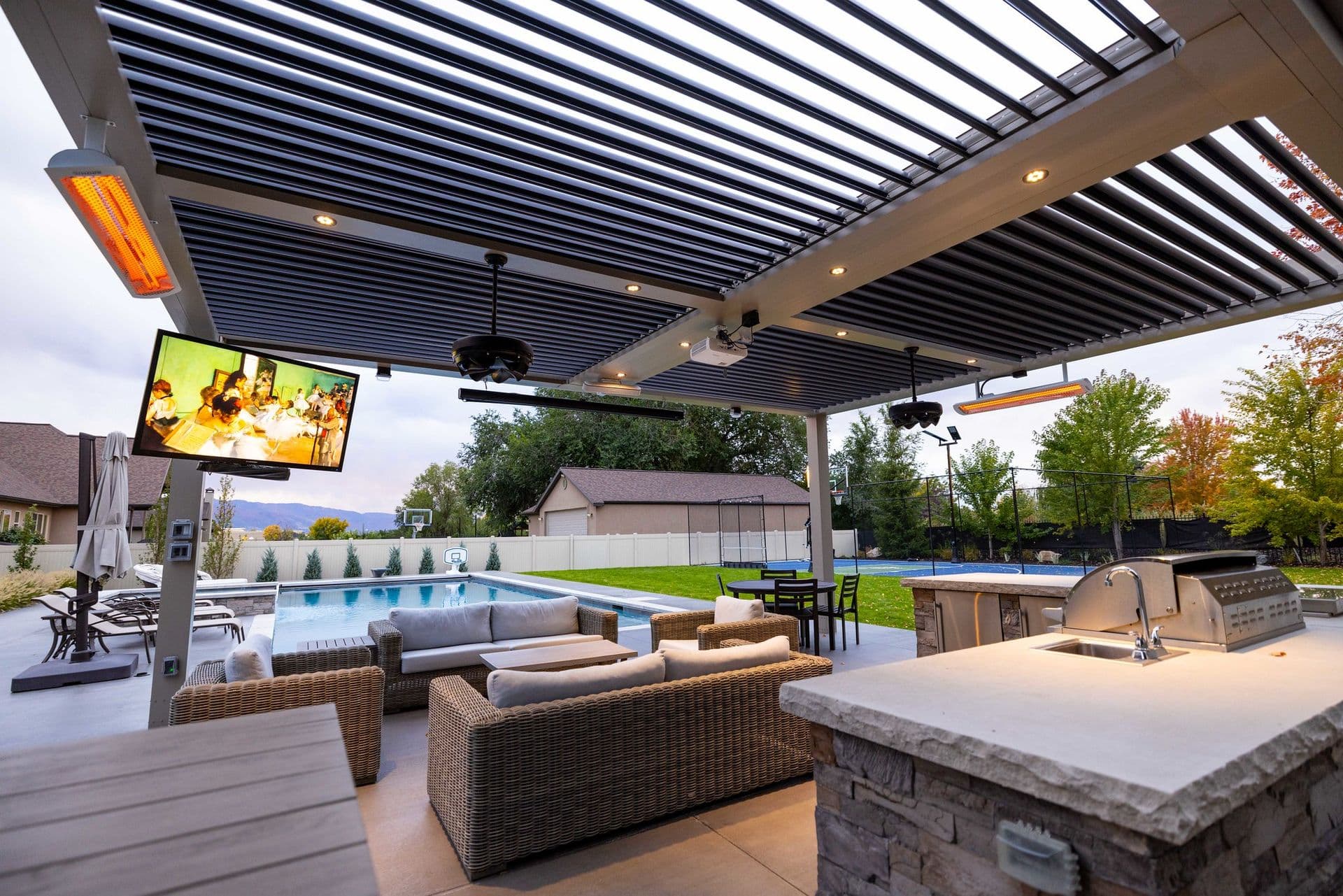 Modern outdoor patio with a louvered roof, cozy wicker seating, TV, grill, and views of a pool and lush garden, creating a luxurious and inviting atmosphere.