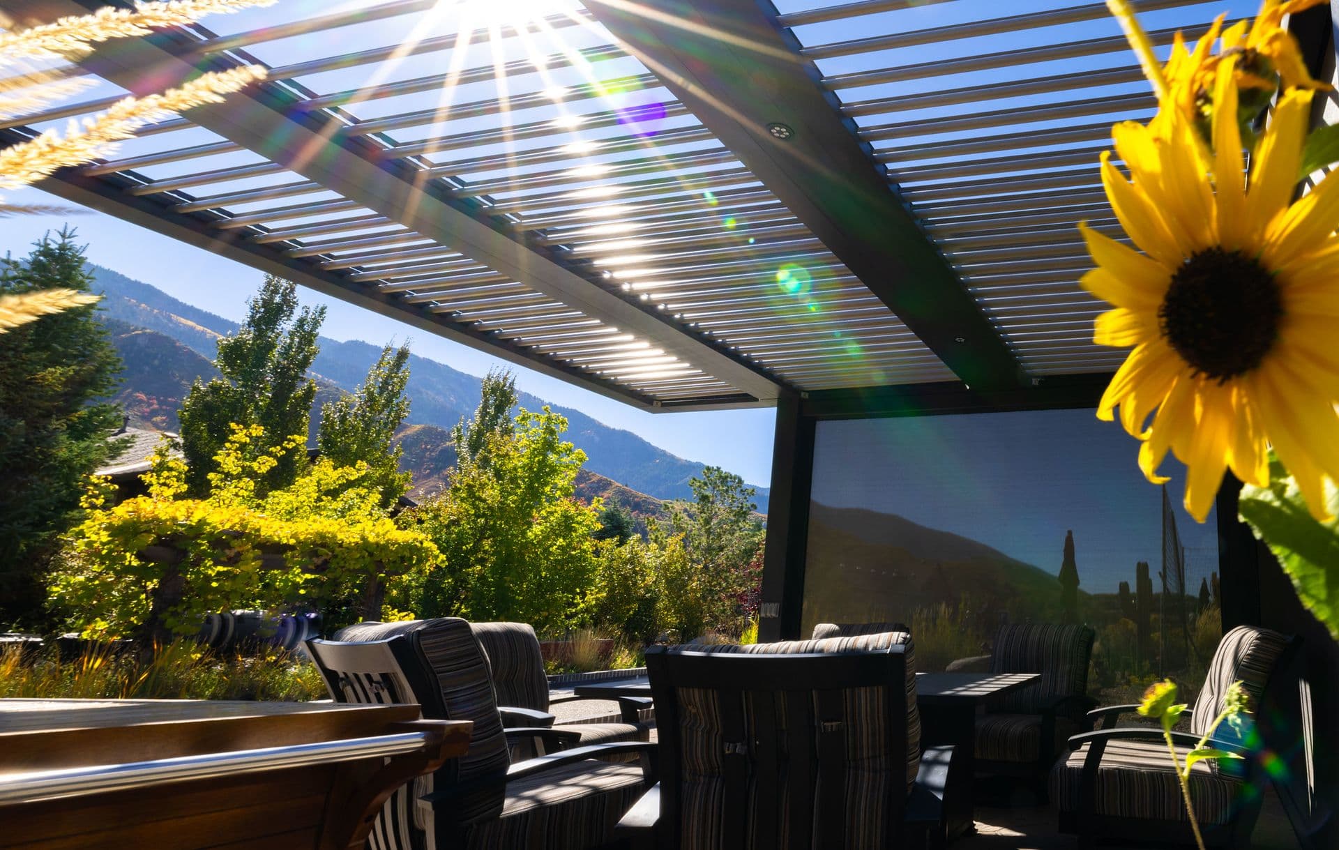 Outdoor patio with striped chairs under a slatted pergola. Sunlight streams through, illuminating a lush garden and sunflower on the right. Bright, serene ambiance.