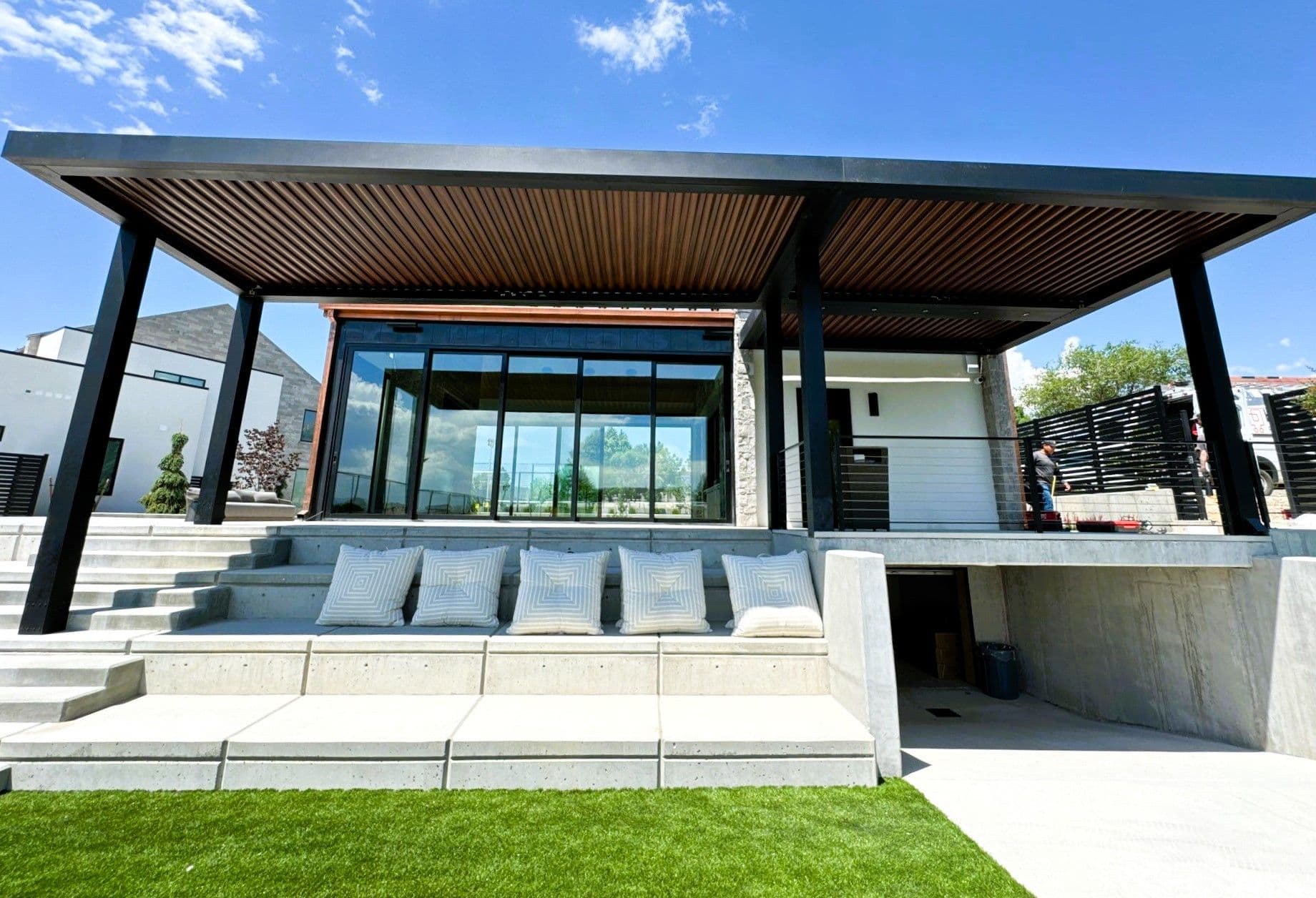Modern home with a sleek pergola and large glass windows. Concrete seating with cushions overlooks a lush green lawn, creating a serene ambiance.