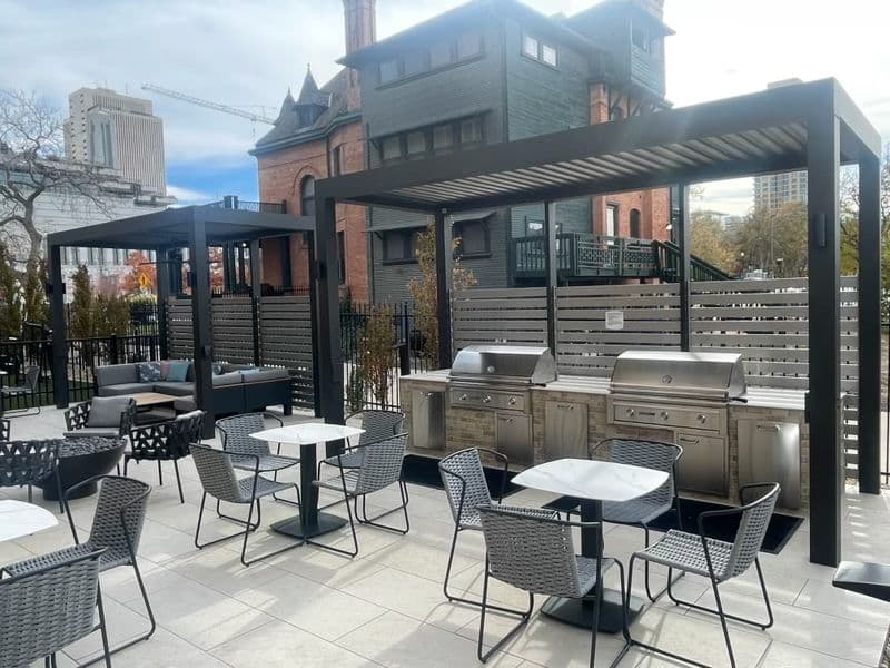 Modern outdoor patio with sleek pergolas, stainless steel grills, and tables with wicker chairs. Brick building and cityscape in the background.