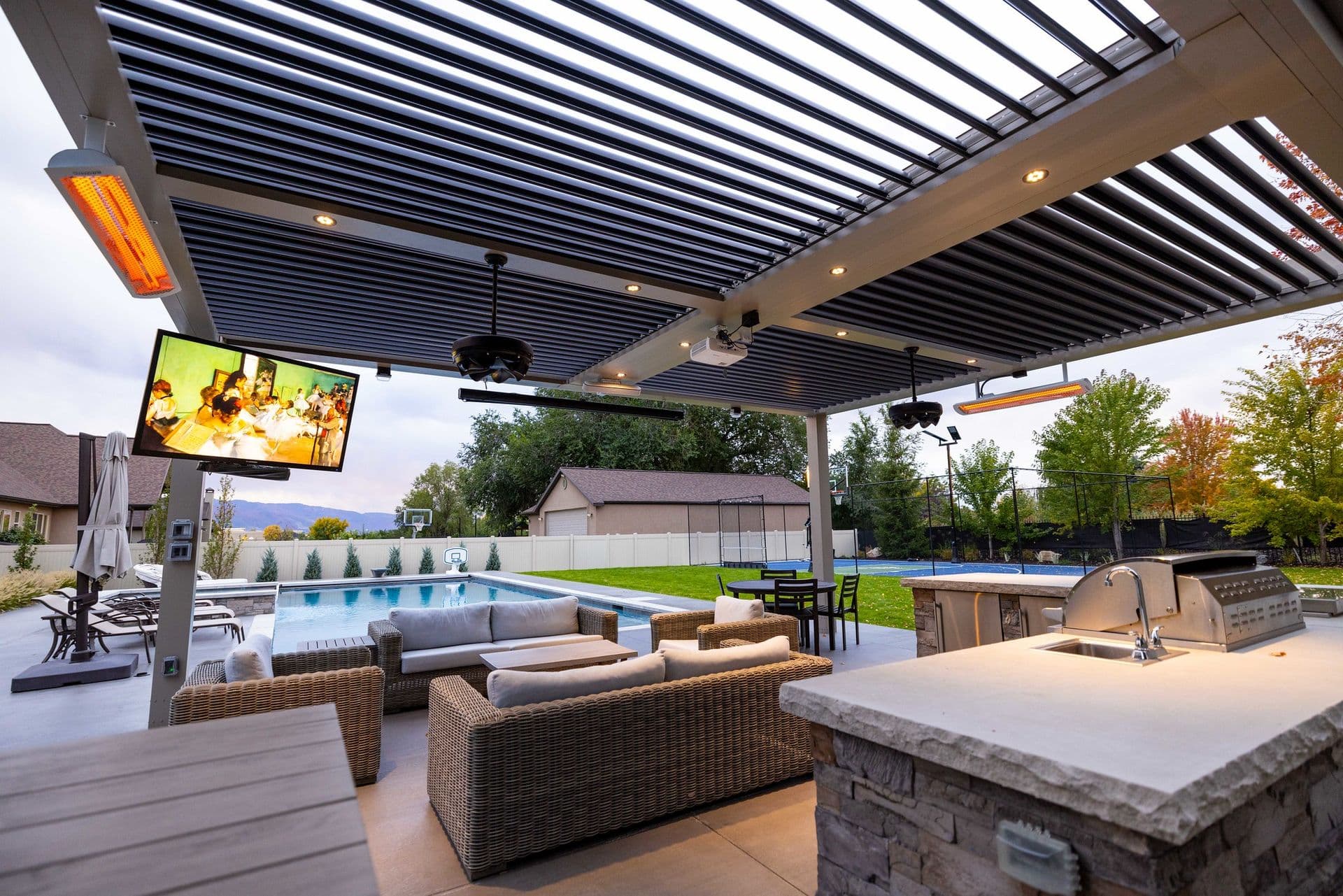 Modern outdoor patio with wicker sofas, TV, and barbecue under a pergola. Overlooks a pool and garden, creating a cozy, inviting ambiance.