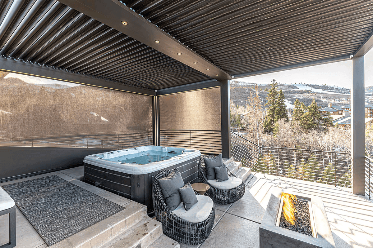 Modern outdoor patio with hot tub under a pergola, cozy chairs, and a fire pit. Overlooks snowy mountains and trees, creating a tranquil setting.