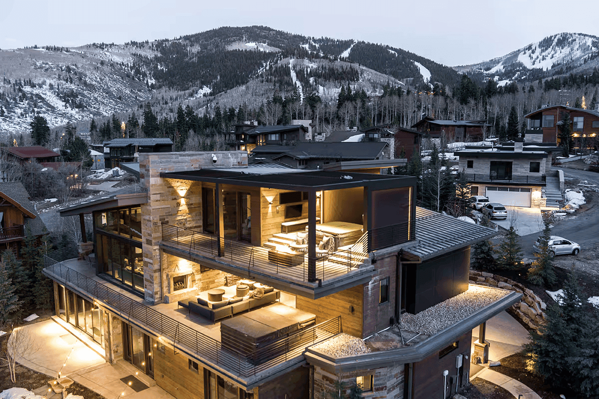 Modern multi-story house with large glass walls, warmly lit interiors, and balconies, set against a backdrop of snow-covered mountains at dusk. Luxurious and serene.