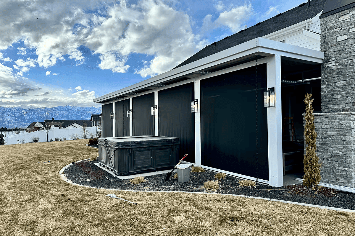 Modern patio with black screens, a covered hot tub, and stone wall on a cloudy day. Snow-capped mountains and residential area in the background.