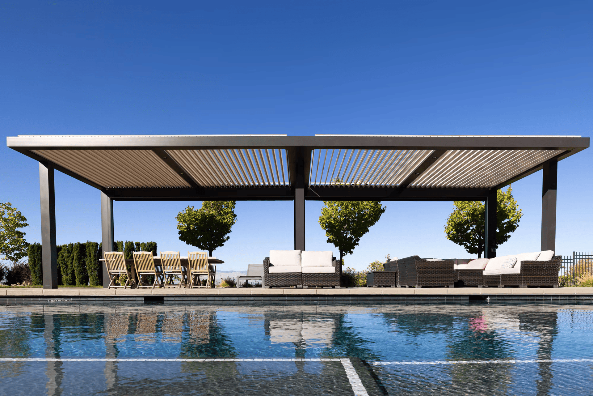 Modern poolside cabana with slatted roof, wicker furniture, and dining chairs. Reflections in the pool, clear blue sky, and neatly spaced trees convey tranquility.