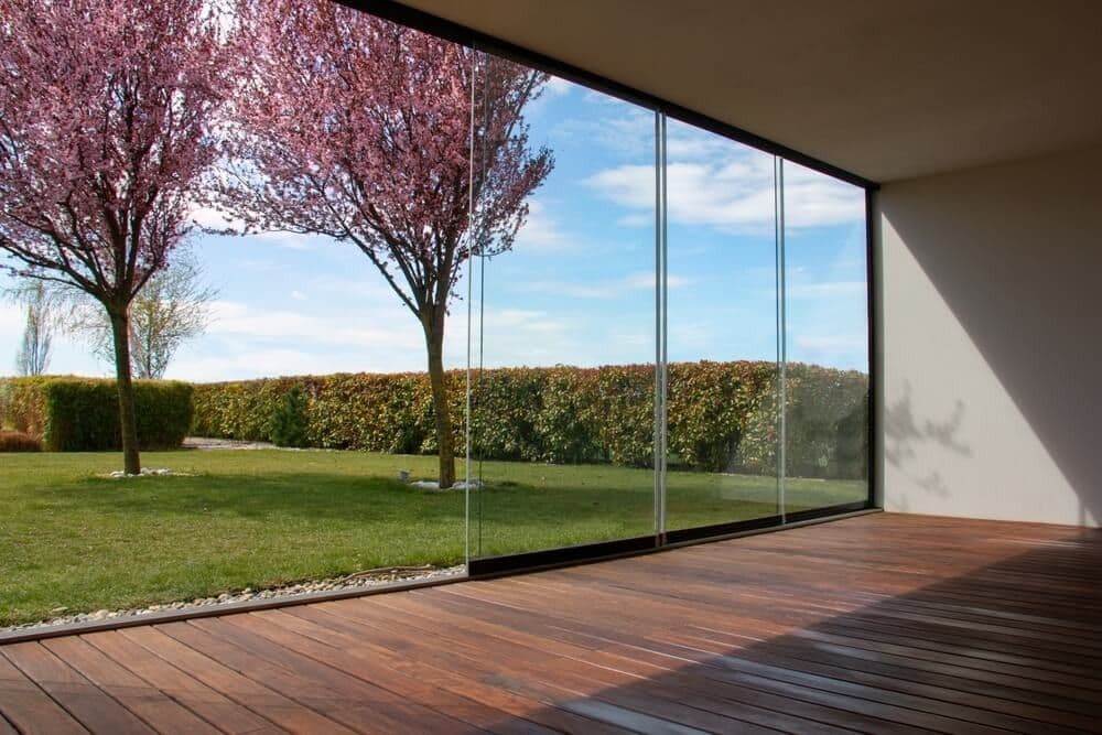 Spacious room with wooden floors and large glass windows. Outside, blooming pink trees and green hedges under a blue sky create a serene view.
