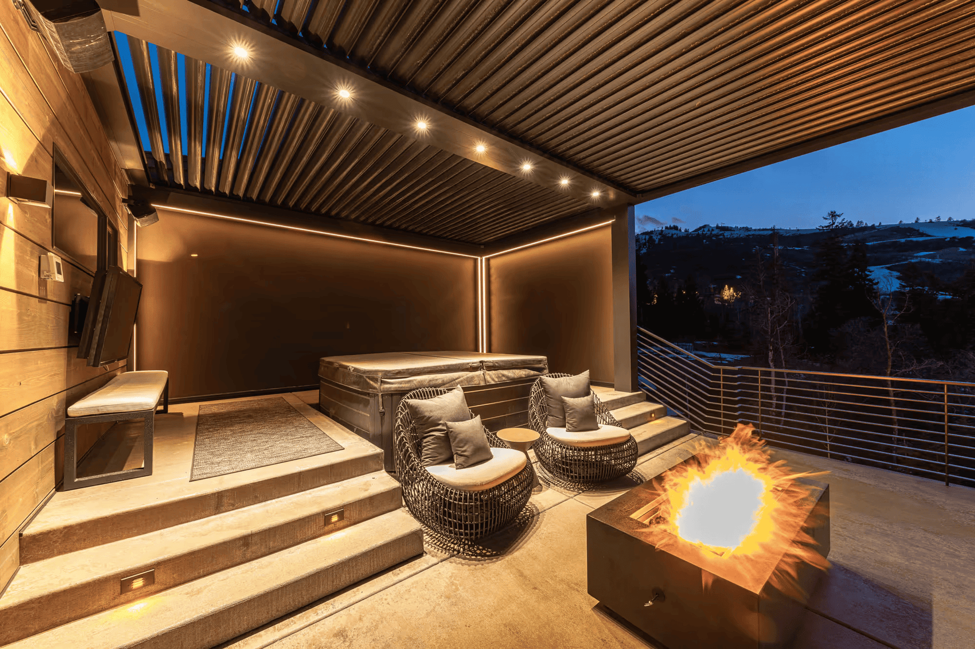 Modern outdoor patio with warm lighting features a hot tub, two cozy chairs, and a blazing fire pit, overlooking a serene evening landscape.