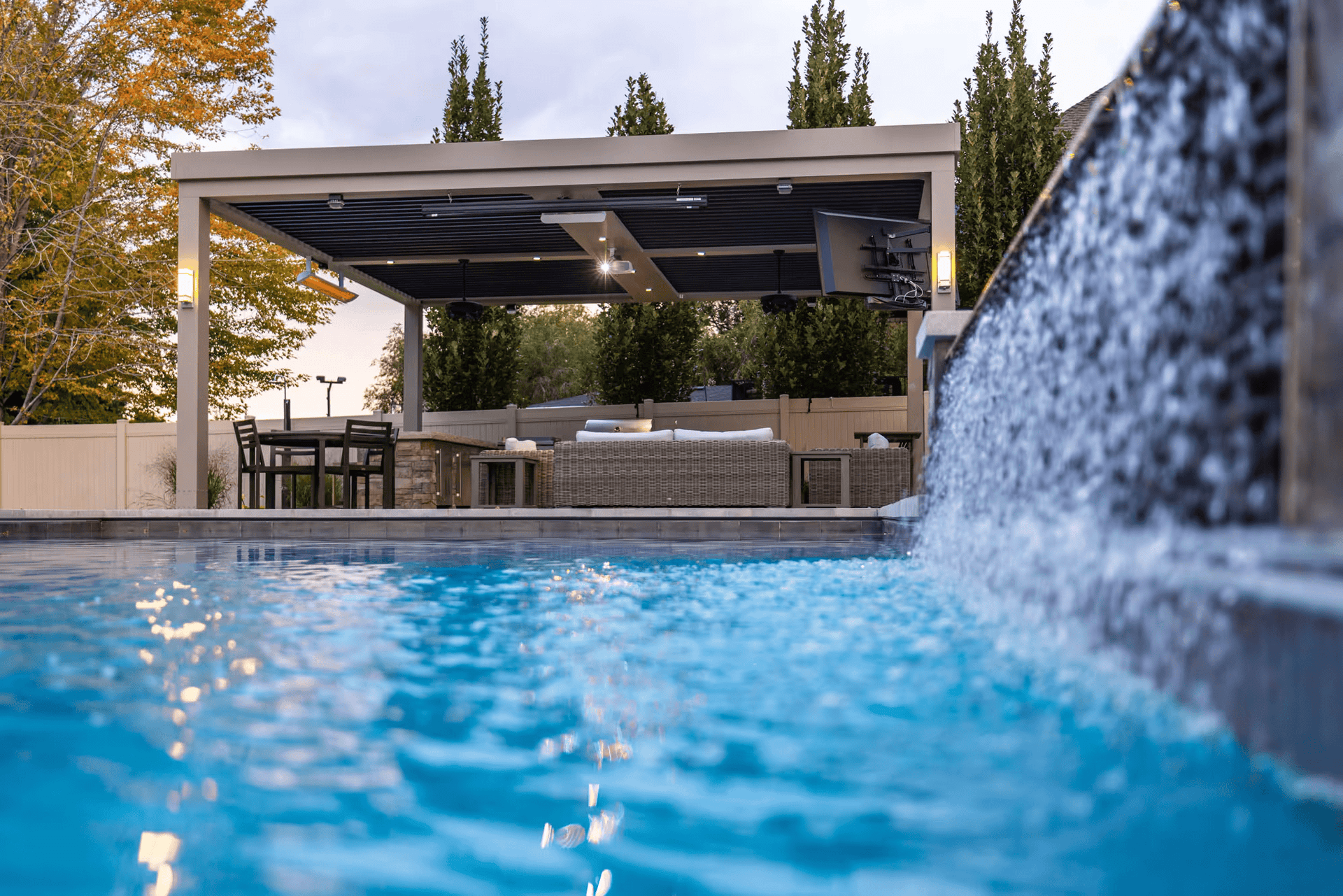 Sparkling pool with waterfall feature in foreground, covered patio with outdoor furniture and TV in background, surrounded by autumn trees. Relaxing atmosphere.