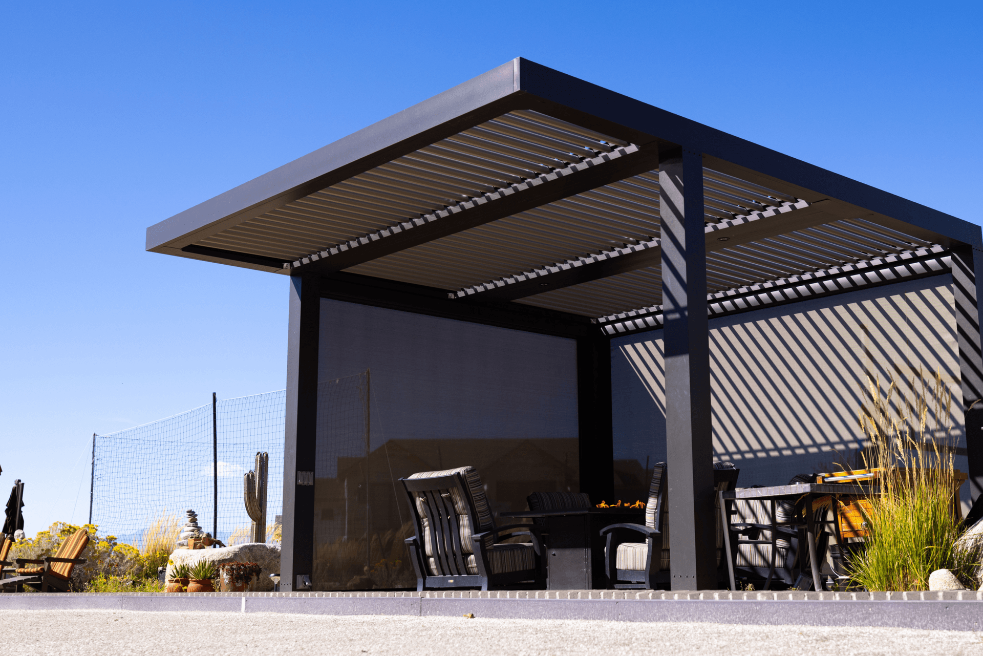 Modern outdoor pergola with sleek black structure casting striped shadows. Beneath are cozy chairs and a fire pit, against a clear blue sky. Relaxed atmosphere.
